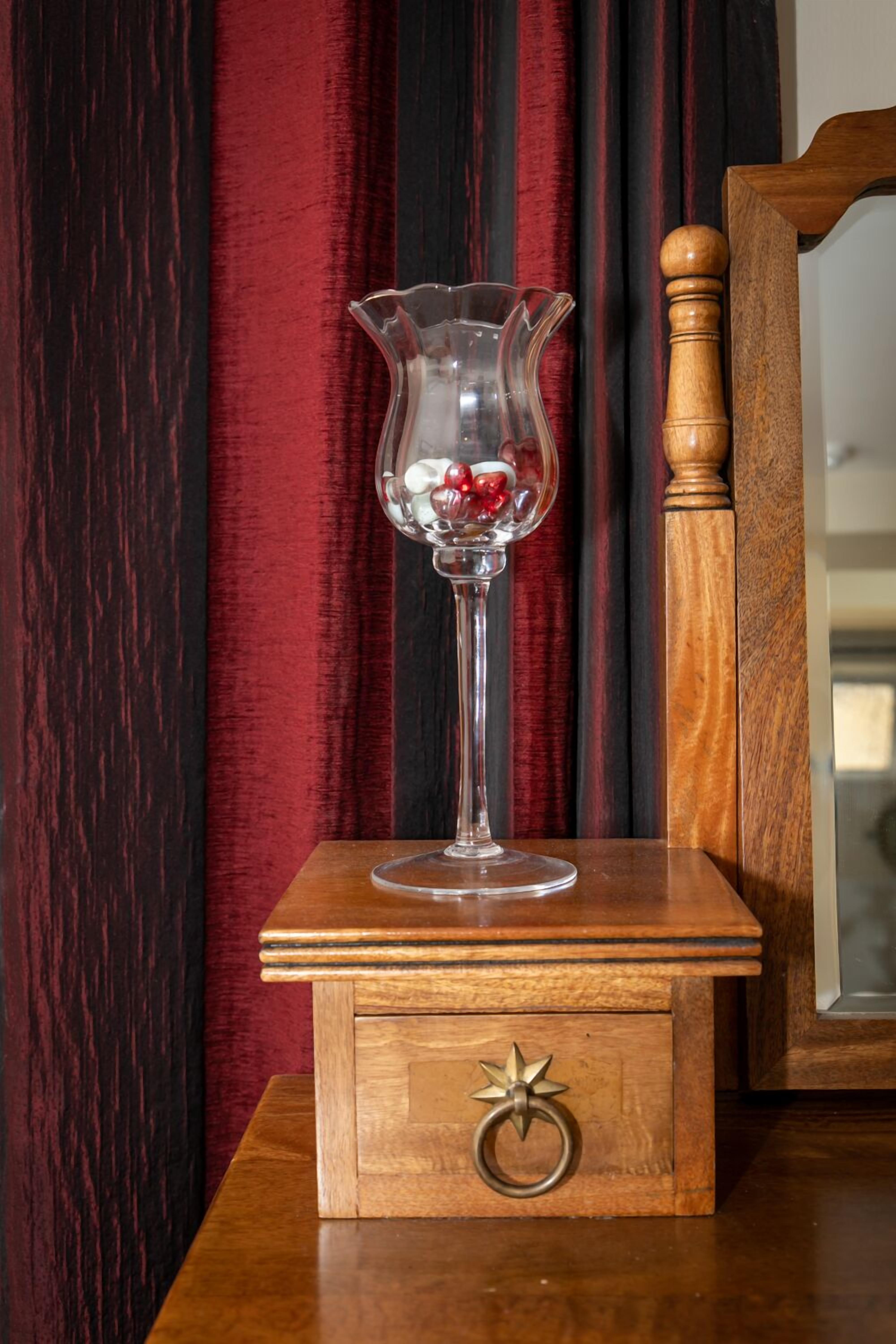 A wooden table with a clear glass vase containing red flowers, set against a backdrop of red curtains and a wooden frame.