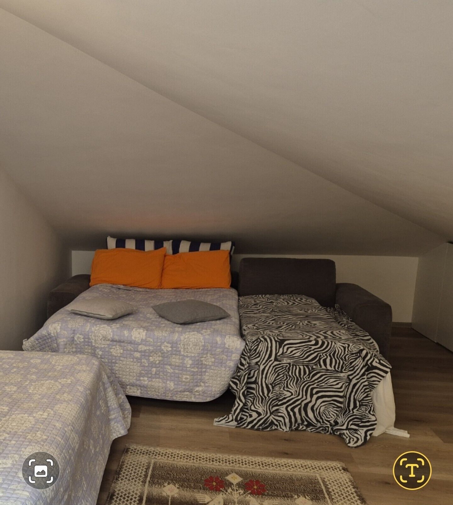 A small, cozy attic bedroom with a sloped ceiling, a bed with patterned bedding, a zebra-print throw, and a small rug with a floral design.