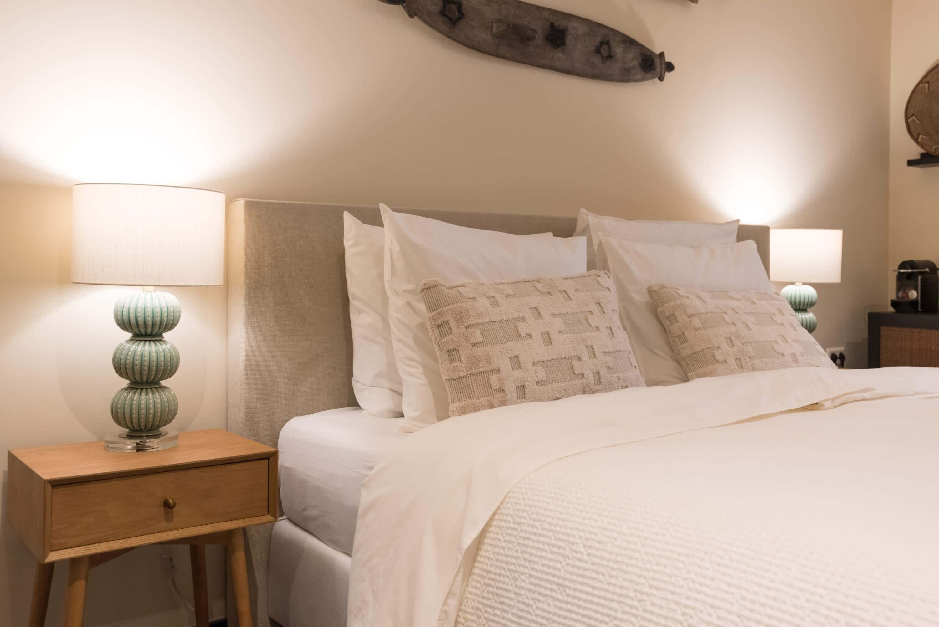 Een netjes opgemaakt bed met witte lakens en twee kussens, een bedlampje en een houten nachtkastje met een lade.