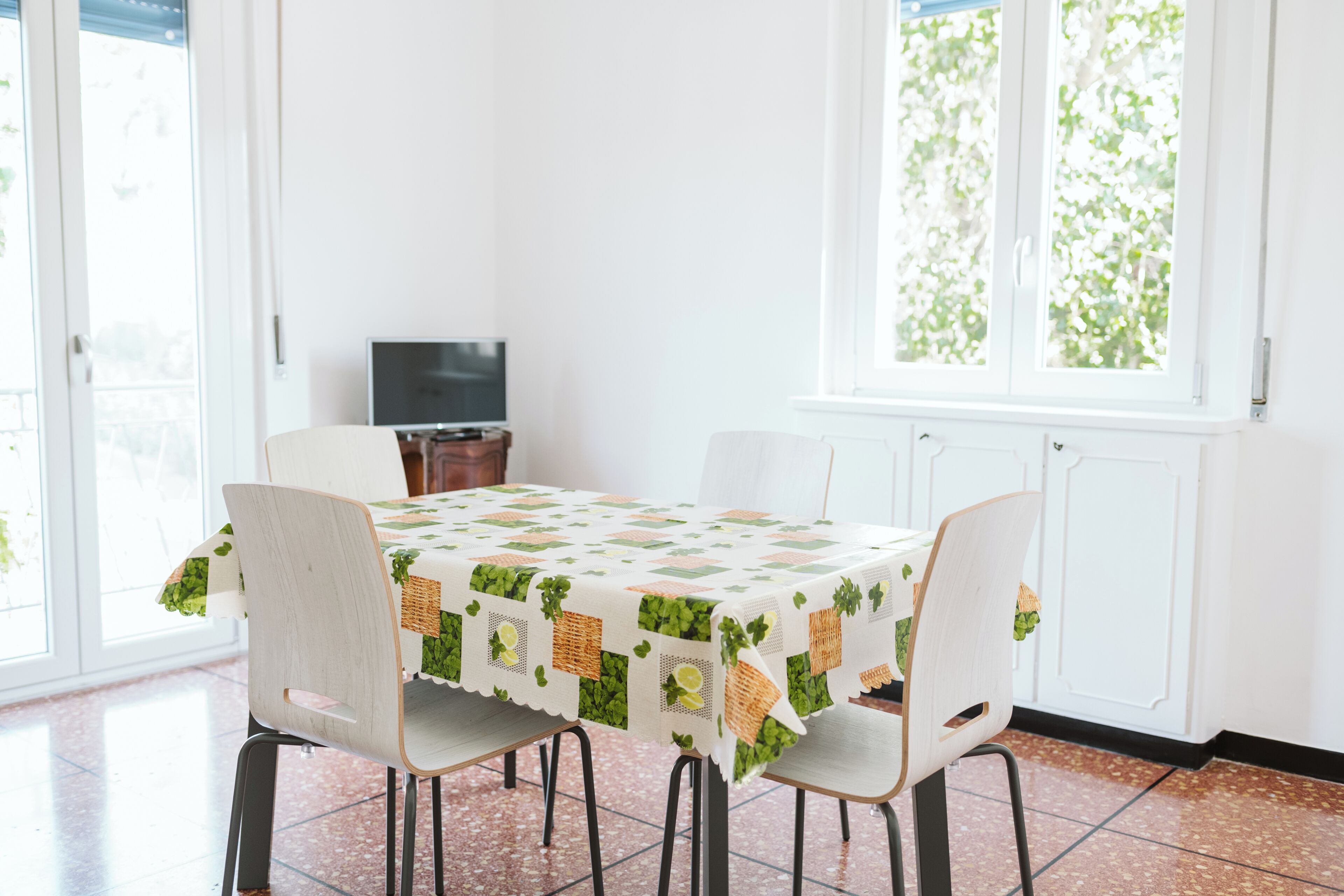 A dining area with a table covered by a patterned tablecloth, four chairs, a television on a stand, and a window with a view of greenery.