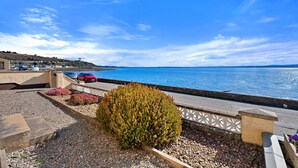 A coastal road with a view of the sea, a parking area, and a landscaped garden with a bush. - The Sea Cottage (Avoch)