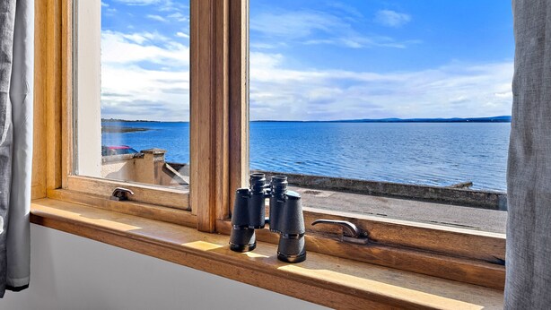 A window with binoculars on a wooden ledge, overlooking a body of water and distant land. - The Sea Cottage (Avoch)