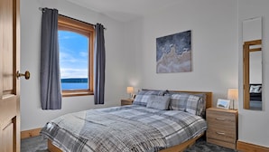 A bedroom with a large bed, a nightstand, a bedside table, a window with curtains, and a painting on the wall. - The Sea Cottage (Avoch)