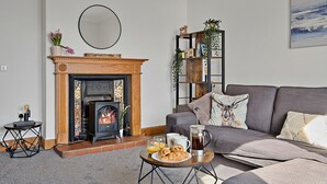 A living room with a fireplace, a sofa, a coffee table with breakfast items, and a bookshelf. - The Sea Cottage (Avoch)