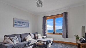 A modern living room with a grey sofa, a small round coffee table, a painting on the wall, and a window with curtains. - The Sea Cottage (Avoch)