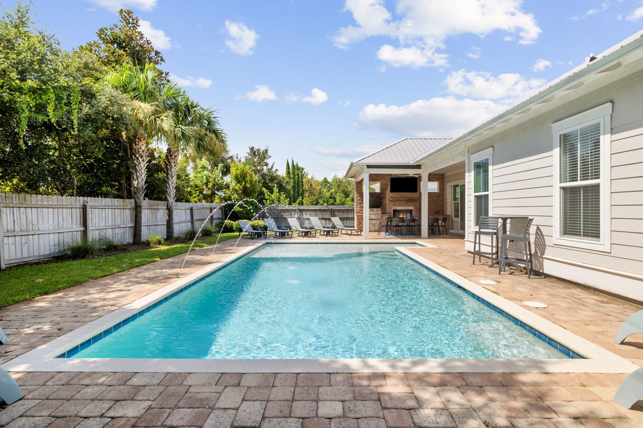 A rectangular swimming pool with a tiled edge, surrounded by a brick patio. There are lounge chairs and a covered patio area with tables and chairs.