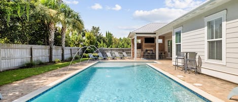Ein rechteckiger Pool mit einer gefliesten Kante, umgeben von einer Pflasterterrasse. Liegestühle und ein überdachter Bereich mit Tischen und Stühlen sind vorhanden.