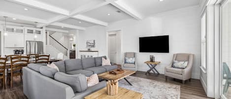 A spacious living room with a gray sectional sofa, a wooden coffee table, and a large flat-screen TV mounted on the wall.