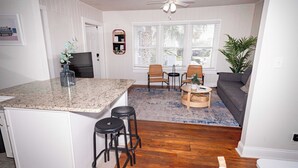A kitchen with a granite countertop, two bar stools, a dining area with a table and chairs, a sofa, and a large window with blinds.