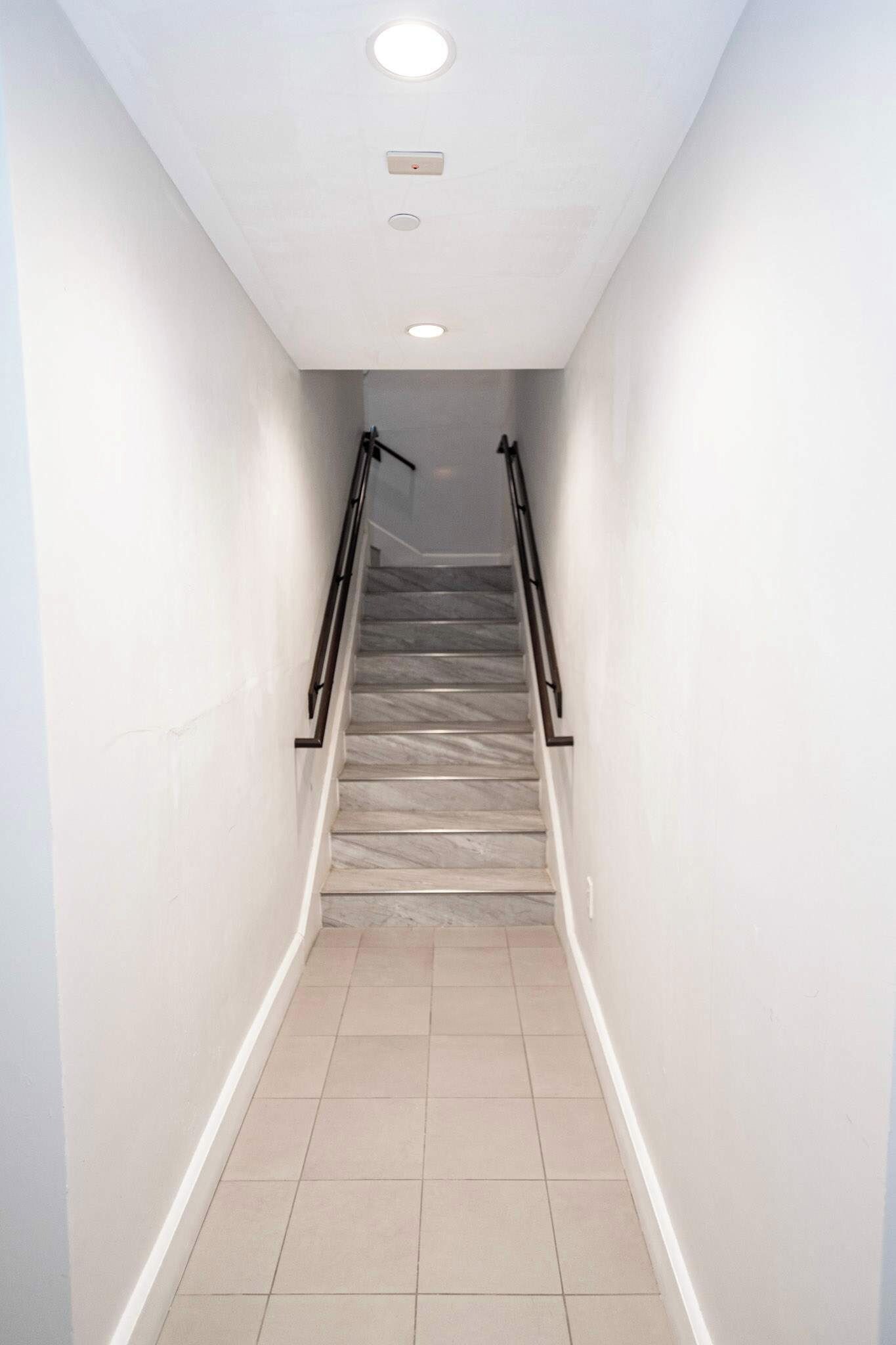 Un corridoio stretto, con pavimento in piastrelle, e una scala che sale, con una ringhiera su un lato e un lampadario al soffitto.