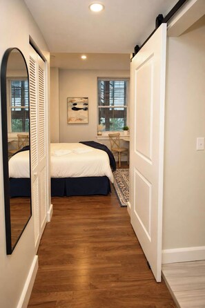 A bedroom with a bed, a chair, a desk, a window, and a mirror.
