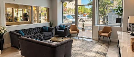 A spacious living area with a sectional sofa, a small round coffee table, and a dining table with chairs. Large windows provide a view of the street and parked cars outside.