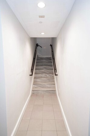 A narrow, tiled hallway with a staircase leading upwards, featuring a handrail on one side and a light fixture on the ceiling.