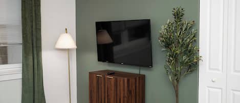Una habitación con una televisión de pantalla plana montada en la pared, una lámpara de pie, un armario de madera y una planta decorativa.