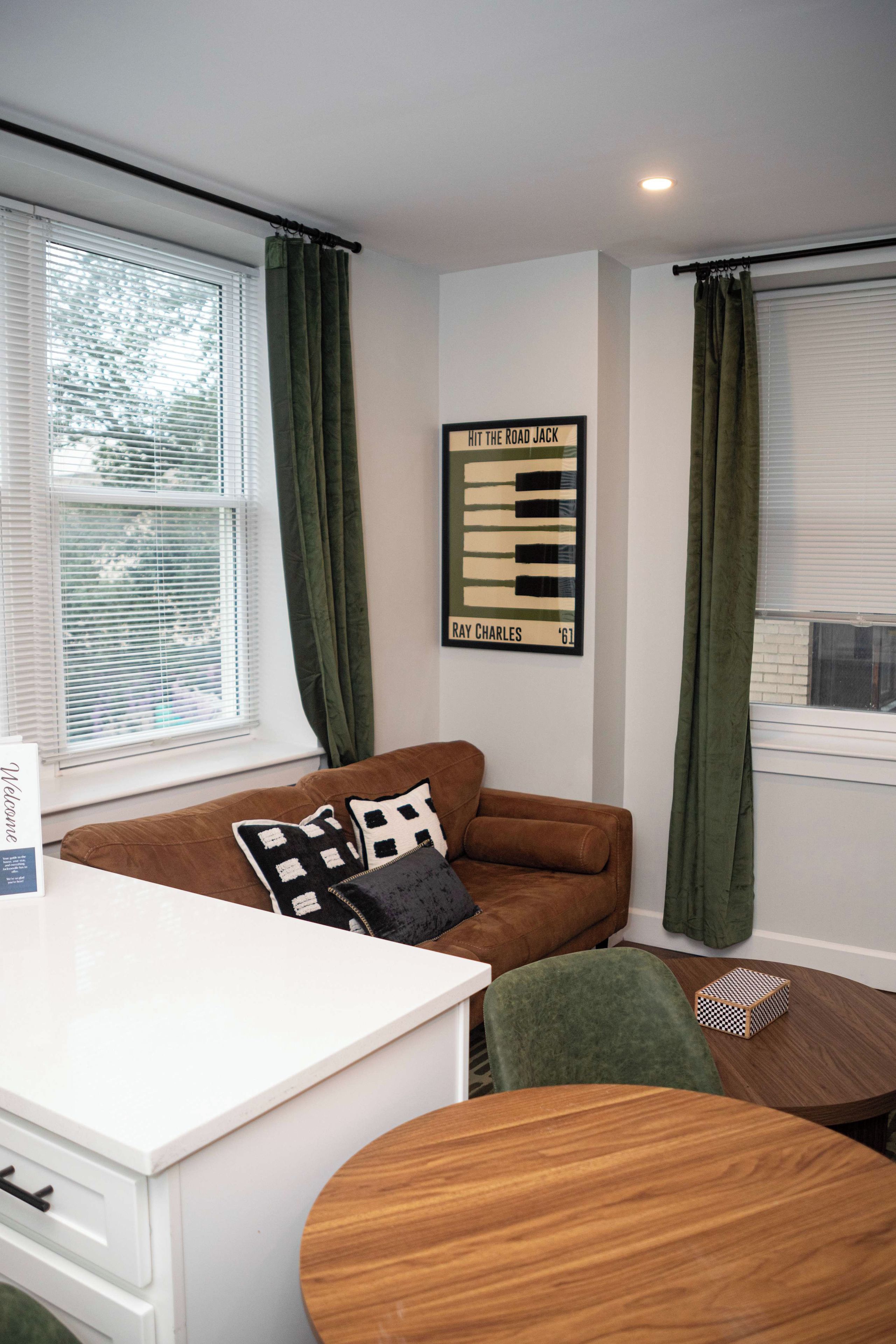 A room with a brown sofa, a white desk, a framed picture on the wall, and a window with green curtains.
