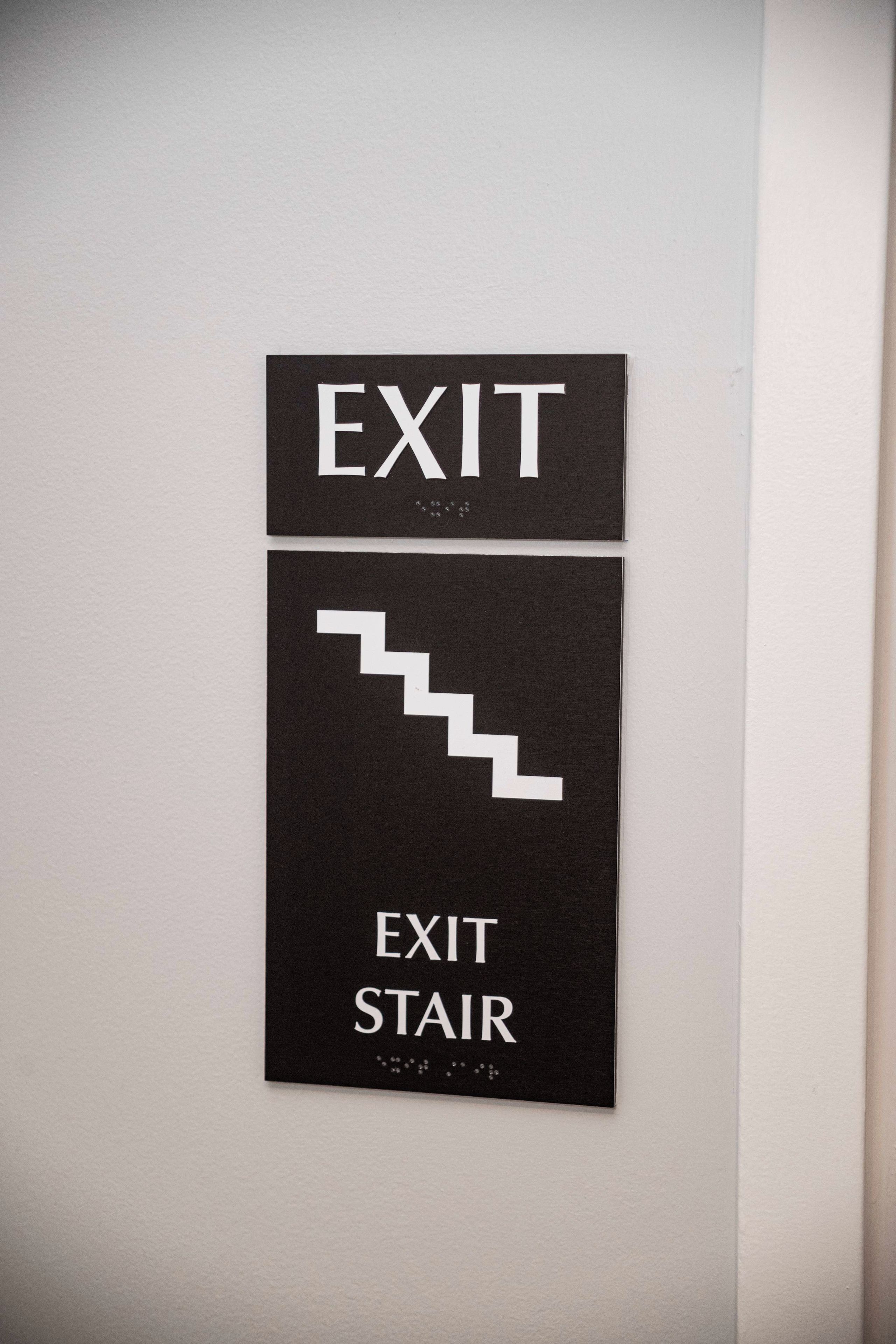 Placa na parede com a palavra 'EXIT' e uma seta apontando para baixo, indicando a direção da saída ou escada.