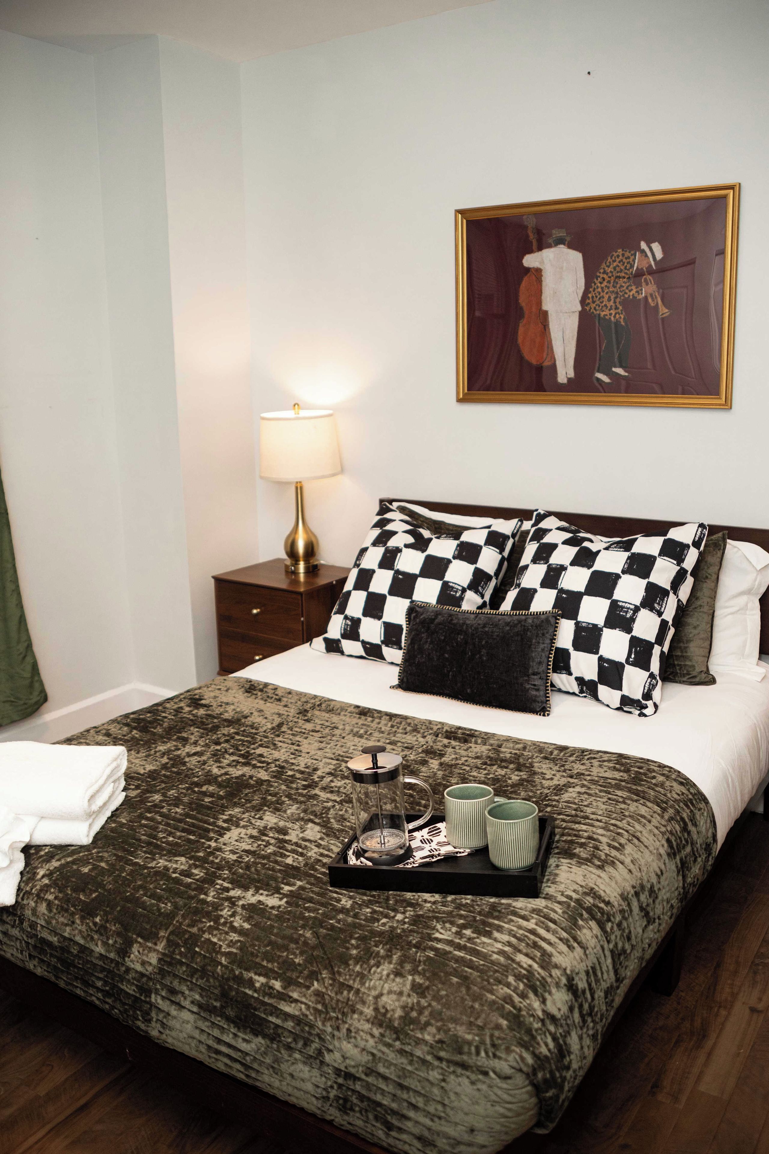 A hotel room with a bed, a nightstand, a lamp, a painting, and a tray with a compass and mugs.