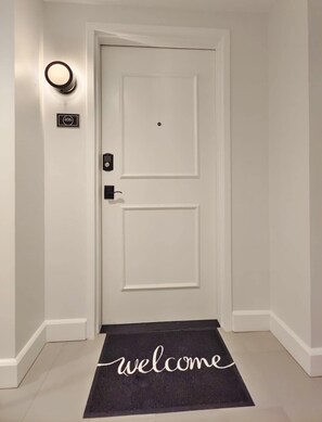 Eine weiße Tür mit einem Zahlenschloss, darüber eine Lampe, und ein Schild mit der Aufschrift '406'. Vor der Tür liegt eine schwarze Fußmatte mit dem Schriftzug 'welcome'.