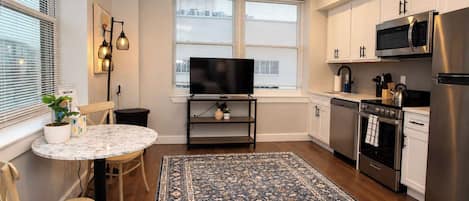 A compact kitchen with a dining area, a flat-screen TV, and a large rug.