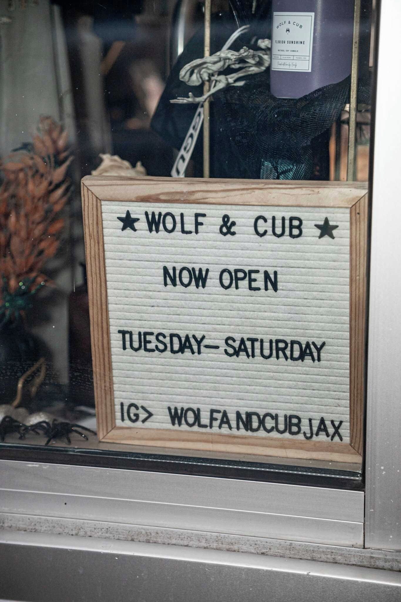 Ein Holzschild mit der Aufschrift 'WOLF & CUB NOW OPEN TUESDAY-SATURDAY' und einem Adresszusatz 'WOLFANDCUBJAX'.