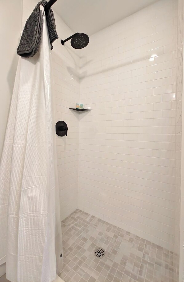 Ducha con paredes de azulejos blancos, cortina de ducha, cabezal de ducha manual y repisa con artículos de aseo.