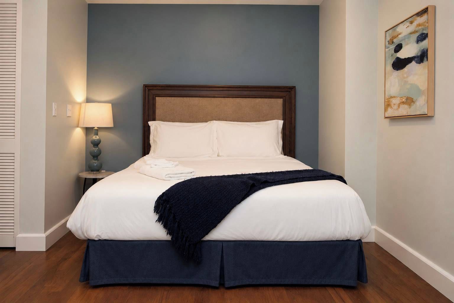 A neatly made bed with white linens and a dark blue skirted bedspread, a bedside lamp, and a painting on the wall.