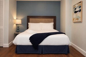 Una cama bien hecha con sábanas blancas y una colcha azul oscuro, una lámpara de noche y un cuadro en la pared.