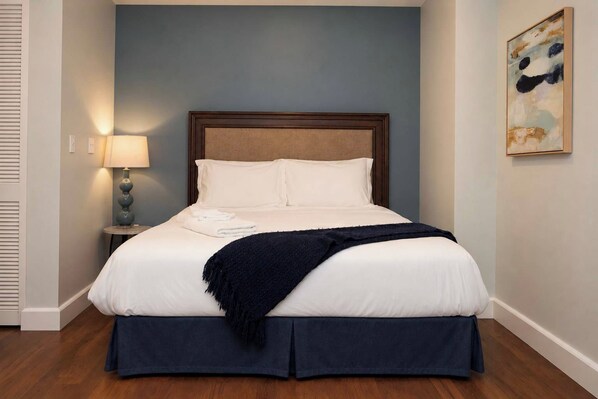 A neatly made bed with white linens and a dark blue skirted bedspread, a bedside lamp, and a painting on the wall.