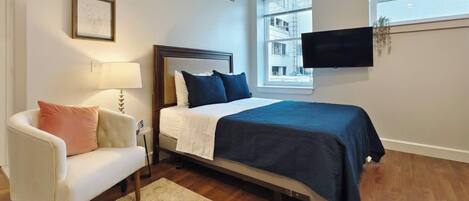 Habitación de hotel con cama, un sillón blanco, una alfombra, televisor montado en la pared y ventana con vistas a edificios.