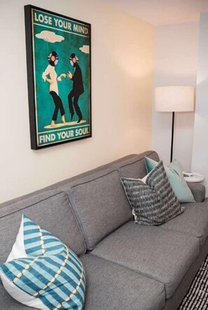 A grey sofa with patterned cushions, a poster on the wall, and a lamp on a side table. - Elegant 1BR King Bed Apts - 1.3 Miles to TIAA Bnk Fld (Jacksonville)