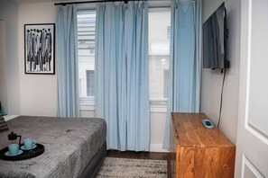 Un dormitorio con cama, una mesita de noche con control remoto, un cuadro en la pared y una ventana con cortinas.