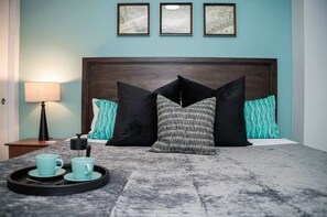A hotel room with a bed, a nightstand, a lamp, and a tray with two cups. - Elegant 1BR King Bed Apts - 1.3 Miles to TIAA Bnk Fld (Jacksonville)