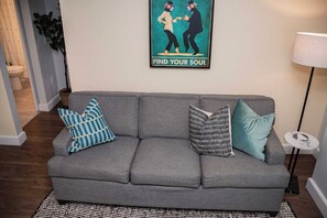Un sofá gris con cojines a rayas y estampados, un cartel enmarcado en la pared y una lámpara a la derecha.