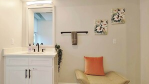 Un baño con tocador blanco, un espejo, un perchero para toallas y dos adornos florales en la pared.
