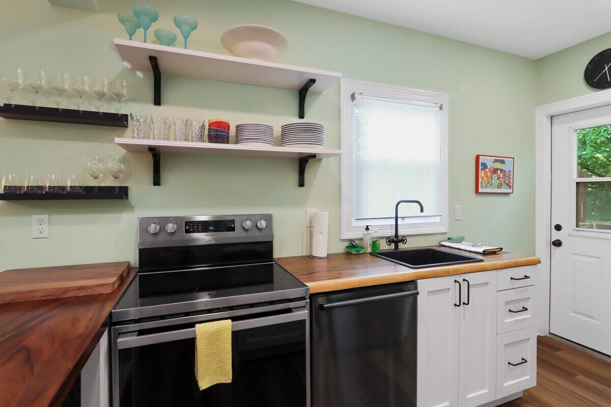 A kitchen with a stove, oven, sink, and white cabinets. There are shelves with dishes and glasses above the stove.