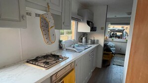 Eine kleine Küche mit weißen Schränken, einem Gasherd und einer Mikrowelle. Eine Holztür führt in einen Wohnbereich mit einer Couch und Blick auf einen parkenden Wagen vor dem Haus.