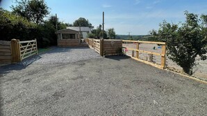 Ein Schotterweg führt zu einem Holztor, einem kleinen Holzgebäude und einem von Bäumen gesäumten, eingezäunten Bereich.