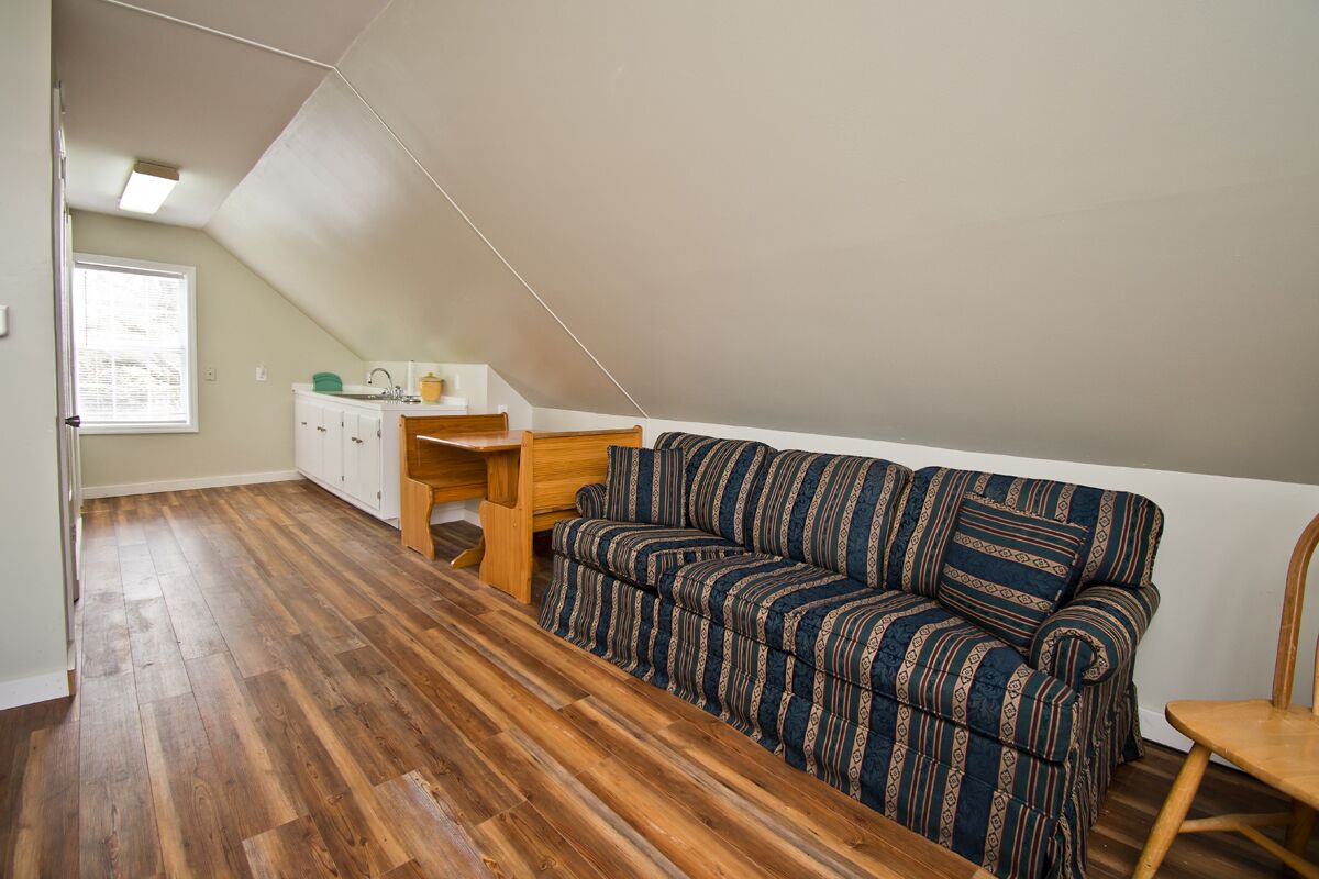 A small, open-plan living space with a wooden floor, a striped sofa, a wooden table, and a kitchenette area.