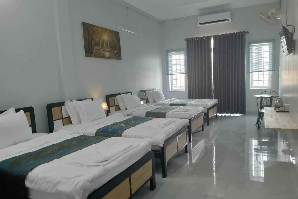 A hotel room with four single beds aligned in a row, a desk with a chair, and a large window with curtains.