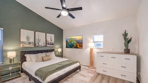 Quarto com cama, mesas de cabeceira, penteadeira, ventoinha no teto, obras de arte e uma janela.