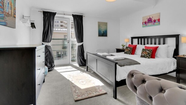 A modern bedroom with a bed, bedside tables, a desk, and a chair. There is a window with curtains, a radiator, and a painting on the wall. - Golden Sunset House Free Parking | By My Getaways (Brighton)