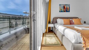 A hotel room with a balcony, a bed with white and orange bedding, a painting on the wall, and a view of the city. - Golden Sunset House Free Parking | By My Getaways (Brighton)