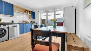 A modern kitchen with blue cabinets, a wooden dining table, and a chair with a red cushion. - Golden Sunset House Free Parking | By My Getaways (Brighton)