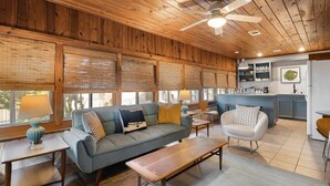 A cozy living room with a sofa, coffee table, and chairs, wooden ceiling, and large windows. - Waterlily Serenity / 2 Bedroom / Creekside (Wimberley)