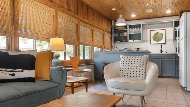 A cozy room with a sofa, a chair, a table, and a kitchen area with shelves and a refrigerator. - Waterlily Serenity / 2 Bedroom / Creekside (Wimberley)