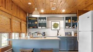 A kitchen with wooden ceiling, a central island, bar stools, and built-in shelves with kitchenware. - Waterlily Serenity / 2 Bedroom / Creekside (Wimberley)
