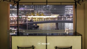 Vista de um quarto com janela que se abre para uma paisagem urbana noturna, com edifícios iluminados, uma ponte e um corpo d'água.