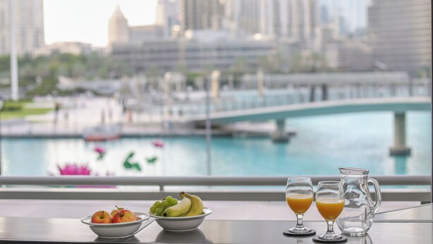 Uma mesa com uma tigela de frutas, dois copos de suco de laranja e um jarro, com vista para uma paisagem urbana que inclui um rio e uma ponte.