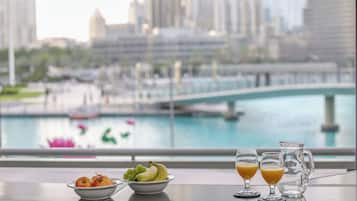 A table with a bowl of fruit, two glasses of orange juice, and a pitcher, overlooking a cityscape with a river and bridge.