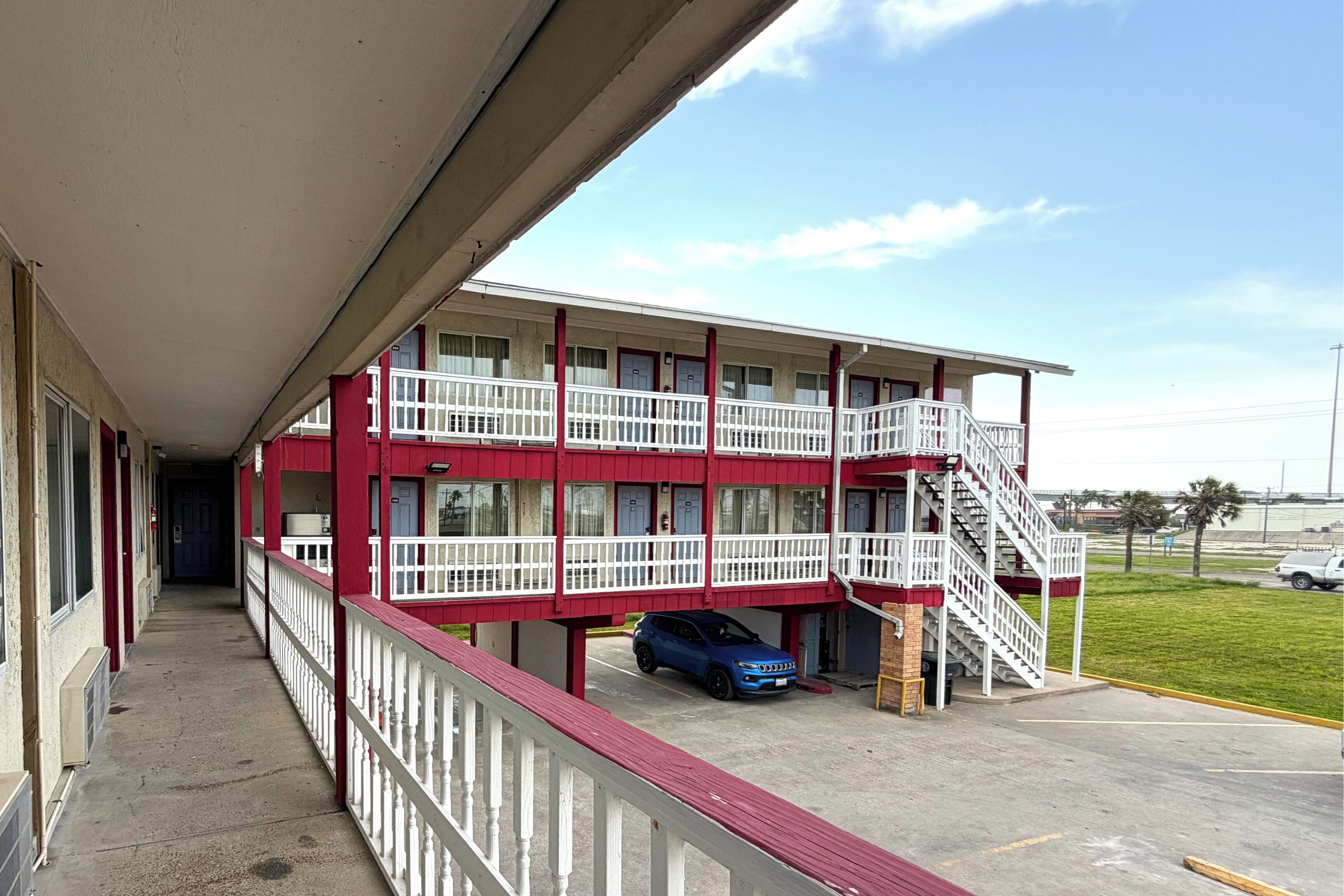 Un motel à plusieurs étages, aux couleurs rouge et blanc, doté de balcons et d’un parking où se trouve une voiture bleue.